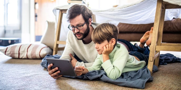 Father and young son lying on the floor under a blanket fort, looking at a tablet together at home