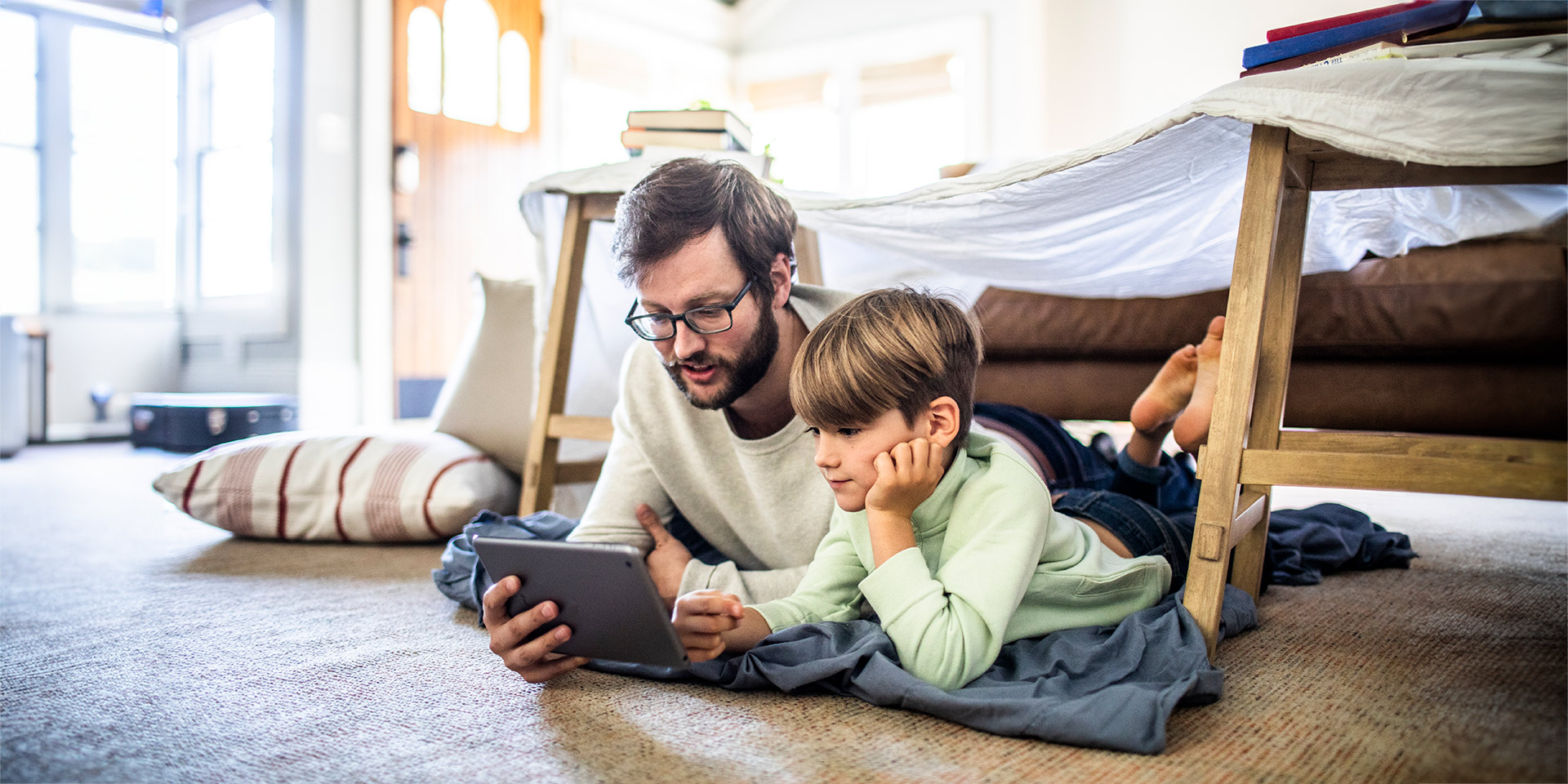 Father and young son lying on the floor under a blanket fort, looking at a tablet together at home